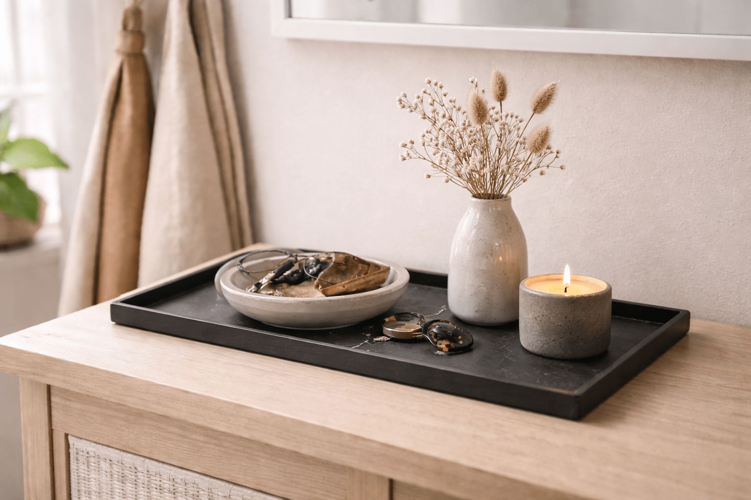 Flaches, rechteckiges Deko-Tablett aus dunklem Metall oder Schiefer auf schmaler Flurkommode mit Keramikschale für Schlüssel, Sonnenbrille, schlichter Vase mit Trockenblumen und kleiner Kerze im Betonhalter, arrangiert im weichen Tageslicht für einen aufgeräumten, einladenden Eingangsbereich.
