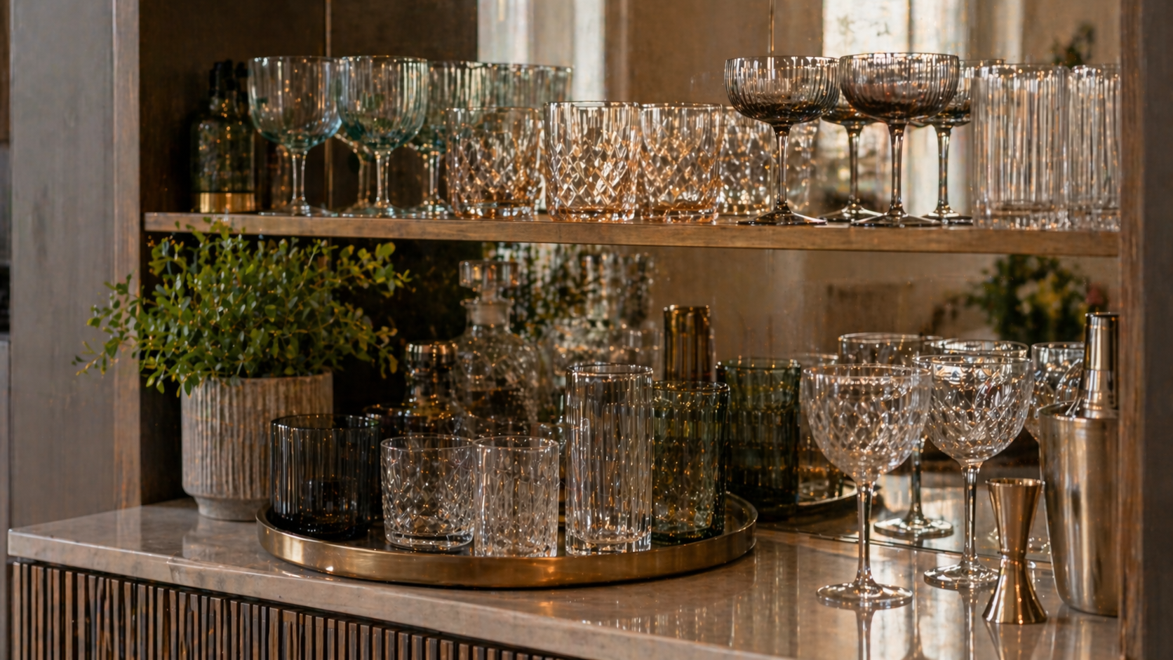 Home Bar mit offenem Holzregal und sorgfältig arrangierten Gläsern in verschiedenen Formen und Höhen, darunter Reliefgläser, farbiges Glas und filigrane Cocktailgläser; das Licht bricht sich in den Strukturen und sorgt für eine warme, wohnliche Atmosphäre.